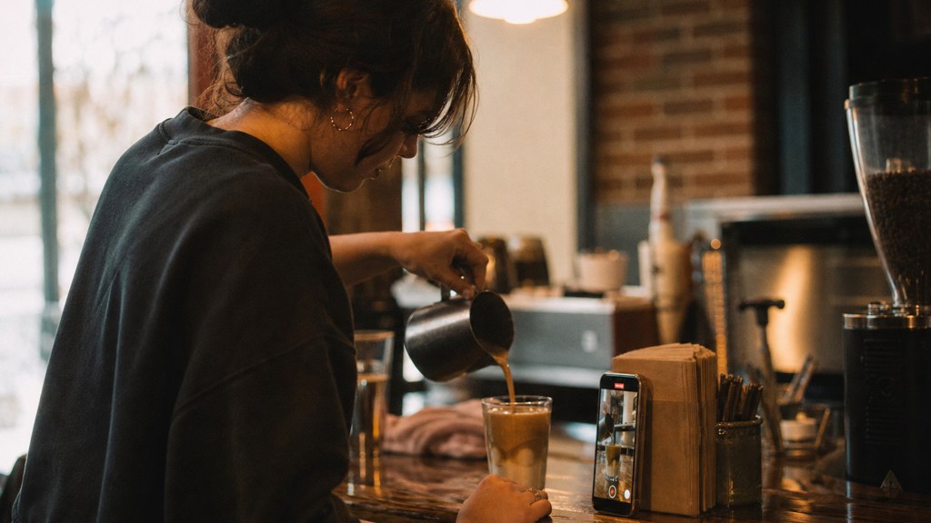 A young cafe worker filming a drink being poured on a phone propped against a napkin holder, photographed in Spokane