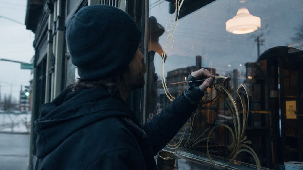 A sign painter finishing gold lettering on a shopfront window from the street side, photographed in Spokane