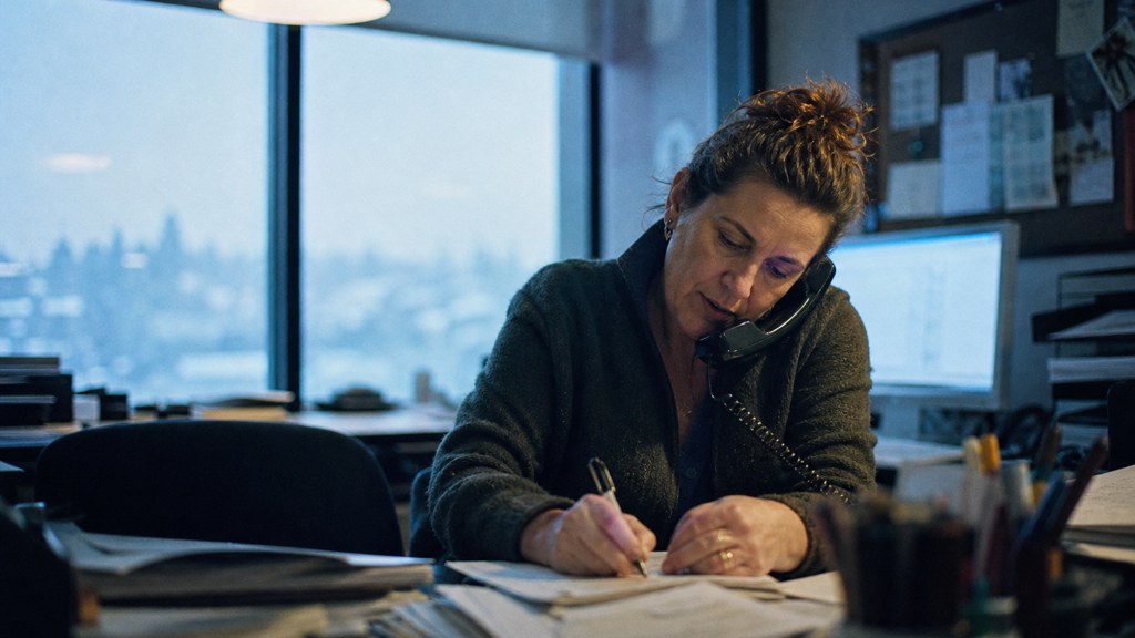 An office manager at a cluttered desk with a landline handset tucked under one ear, photographed in Spokane