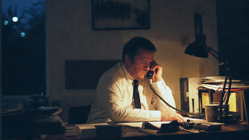 Night-duty director taking a call by a single desk lamp in a quiet office, paperwork and a cold cup of tea beside the phone