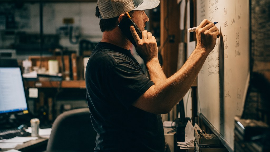 Owner of a small overhead door company taking a call in a cluttered workshop office while writing a job on a scheduling whiteboard