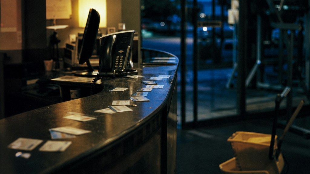 Empty front desk of a fitness club at closing time, membership cards scattered, blue dusk through the glass front