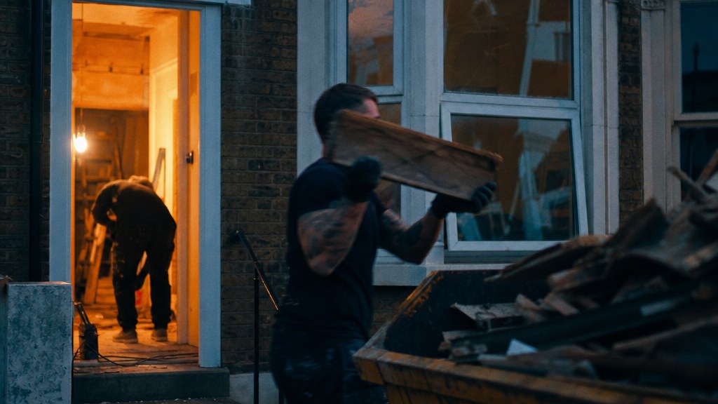 Renovation crew loading offcuts into a skip outside a terraced house at dusk, work lamp glowing amber in the doorway