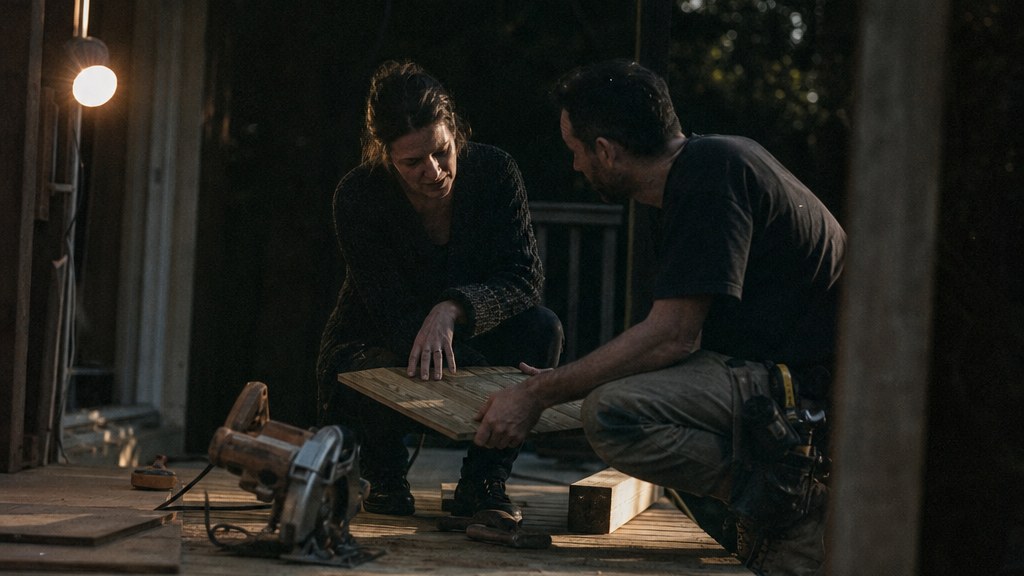 Household owner and fitter crouched on a half-finished deck comparing a sample board in hard afternoon side light