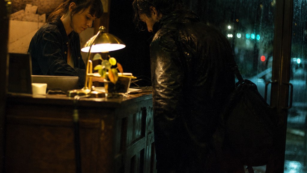 Night receptionist checking in a soaked late arrival in an independent hotel lobby at 2am, single desk lamp, rain on the glass doors