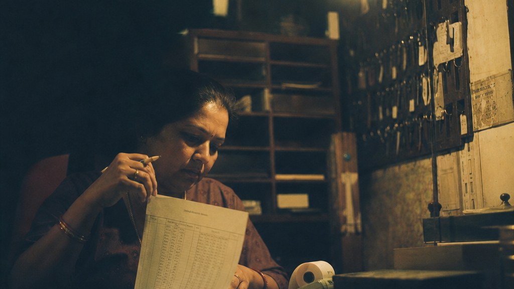 Hotel owner at a back-office desk under a bare bulb reconciling a paper occupancy sheet, keys and a till roll beside her