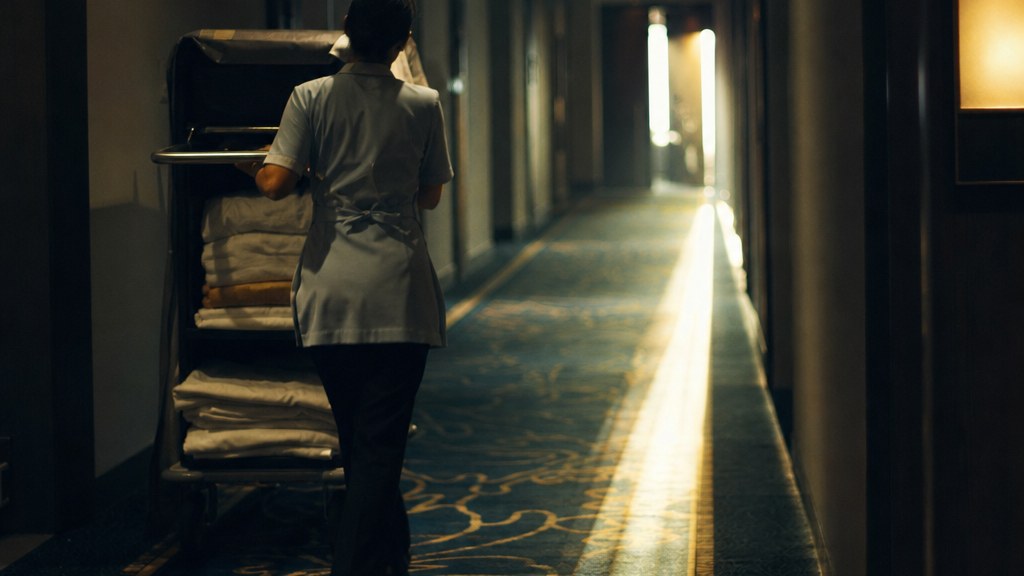 Housekeeper pushing a linen trolley down a dim corridor at first light, a stripe of sun across patterned carpet