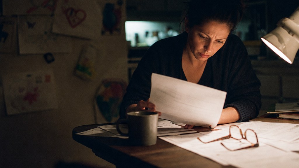 A woman at a kitchen table late at night comparing printed policy pages by lamp light