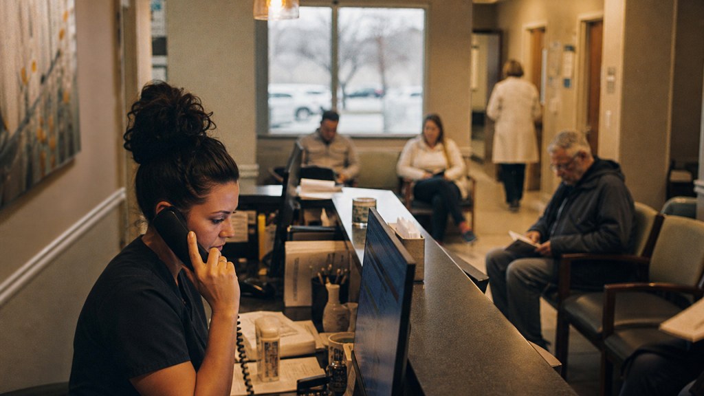 A clinic receptionist taking a phone call at a busy front desk with patients waiting, photographed in Tulsa