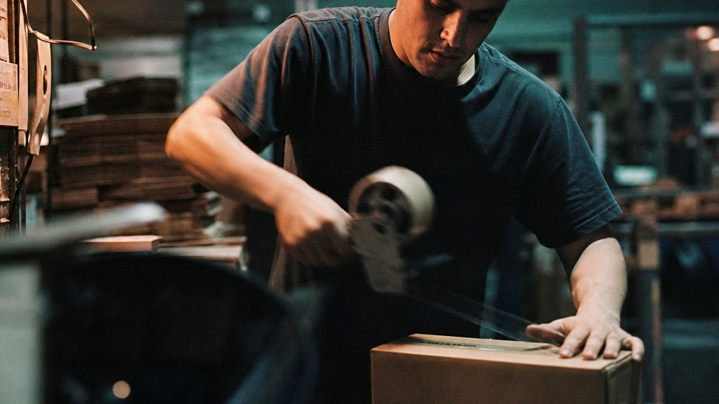 Packer taping the last carton of a night shift under a hanging bulb, tape gun mid-pull