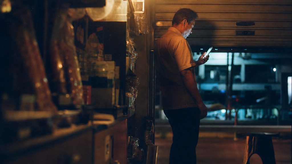 Shop owner alone in a closed store after midnight lit only by the glow of a phone, shutters half down