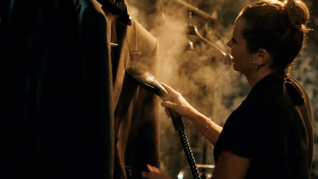 Stylist steaming a jacket on a rail backstage under a bare bulb, steam catching the light