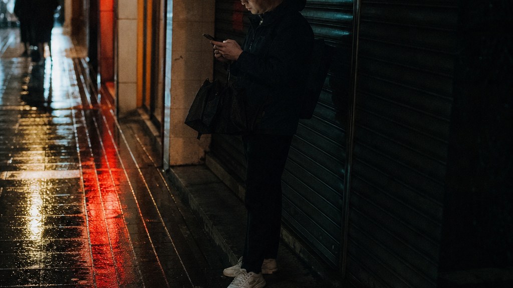 Shopper standing in the rain outside a shuttered shop with a thumb on a phone screen, neon reflecting on wet pavement