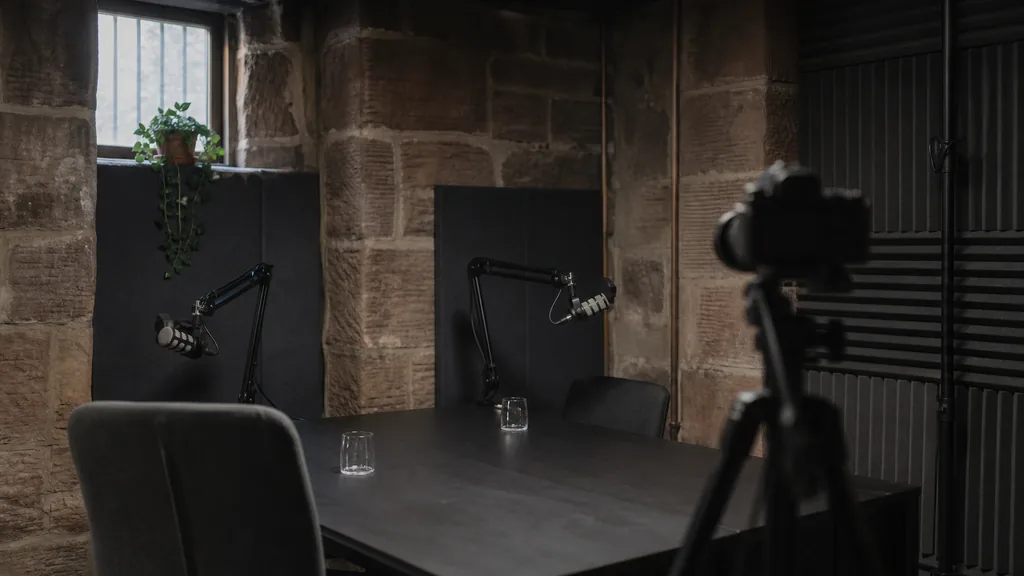 Editorial photograph of an empty podcast studio in a Glasgow sandstone basement conversion with two microphones on boom arms, a camera on a tripod and dark acoustic panels, lit by cool soft northern daylight