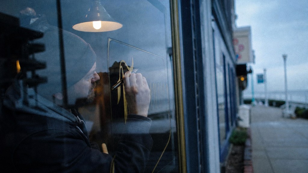 A sign painter finishing gold lettering on a shopfront window from the street side, photographed in Virginia Beach