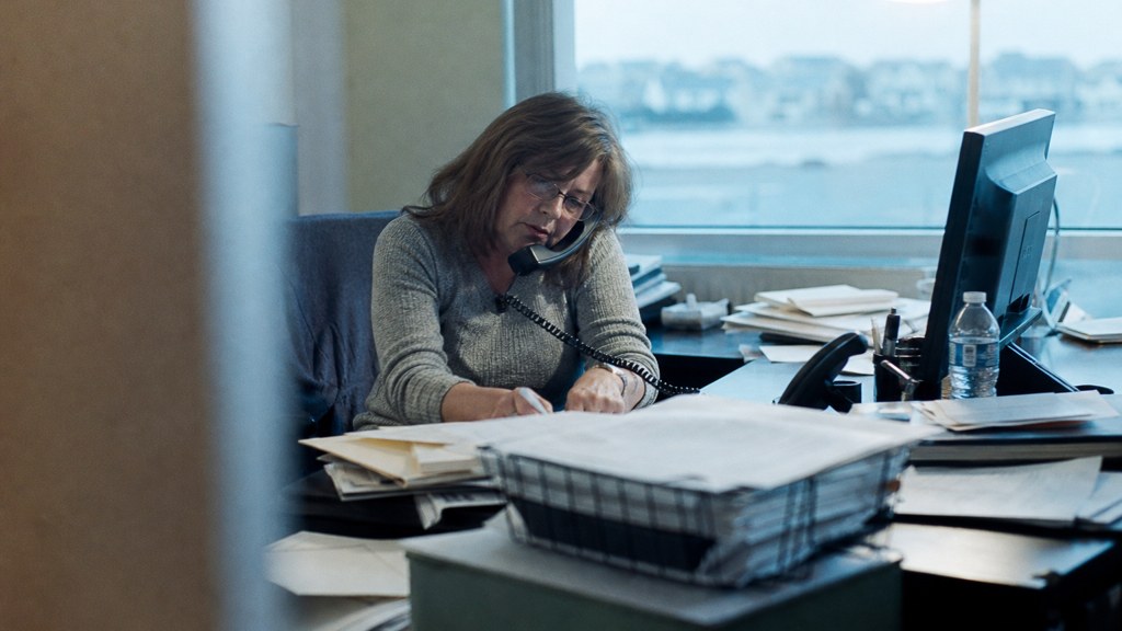 An office manager at a cluttered desk with a landline handset tucked under one ear, photographed in Virginia Beach