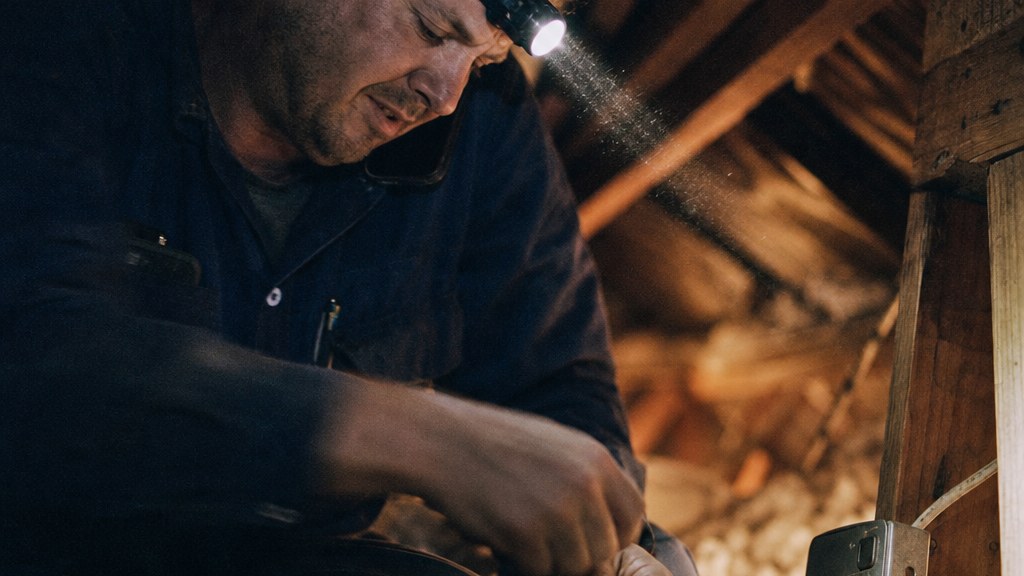 Electrician in an attic crawlspace splicing a wire while taking a booking call on his phone