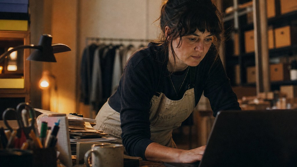 A small-business owner in an apron leaning over a laptop on a cluttered back-office desk late in the day, photographed in Wichita