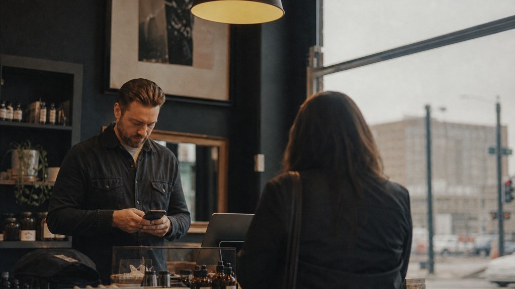 A shop manager standing behind the counter checking a phone while a customer waits at the till, photographed in Wichita