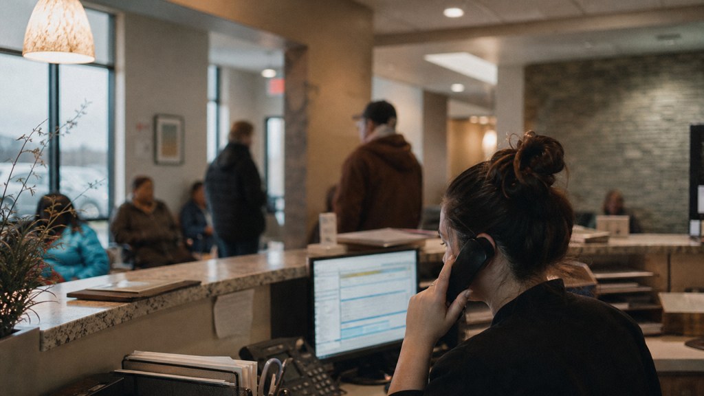 A clinic receptionist taking a phone call at a busy front desk with patients waiting, photographed in Wichita