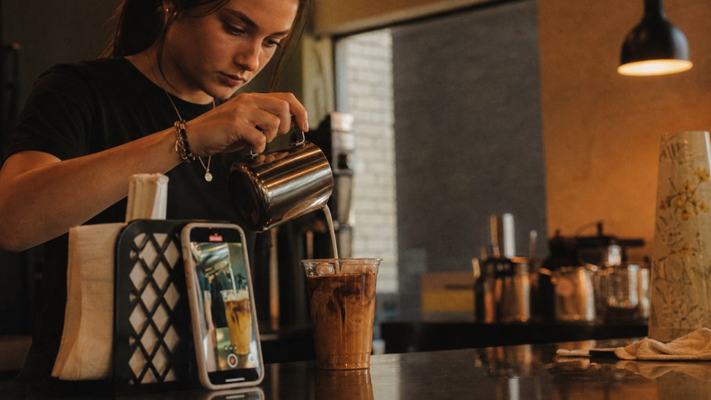 A young cafe worker filming a drink being poured on a phone propped against a napkin holder, photographed in Wichita