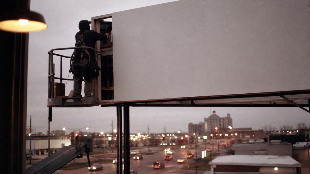 A crew member on a lift installing a blank billboard panel above a busy street, photographed in Wichita