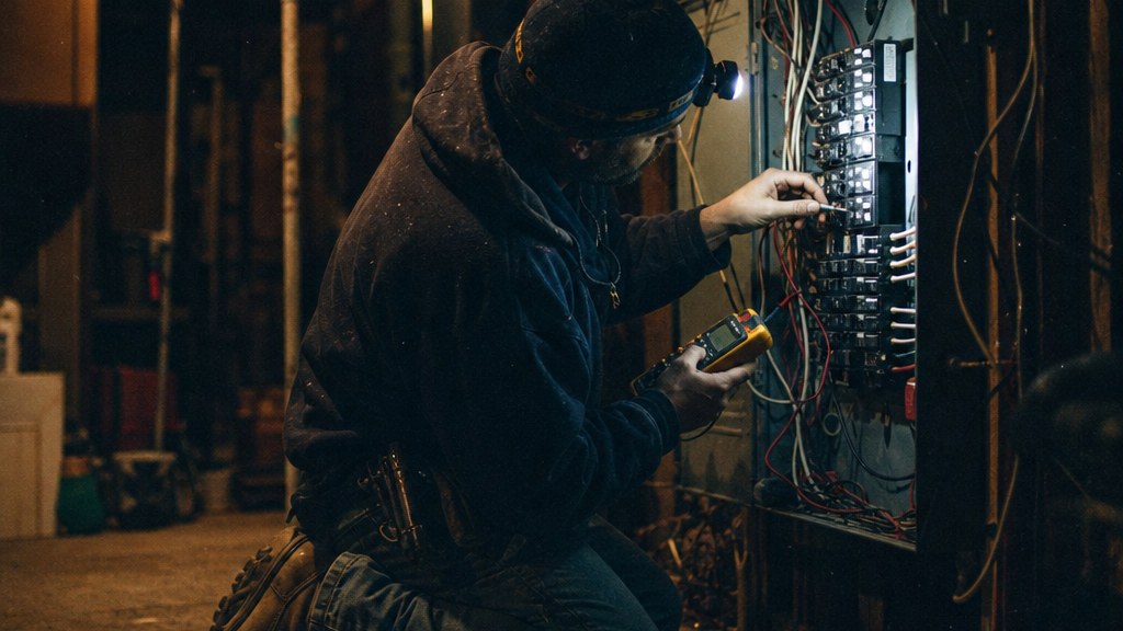 Electrician kneeling in an unlit basement testing an open breaker panel with a multimeter by headlamp