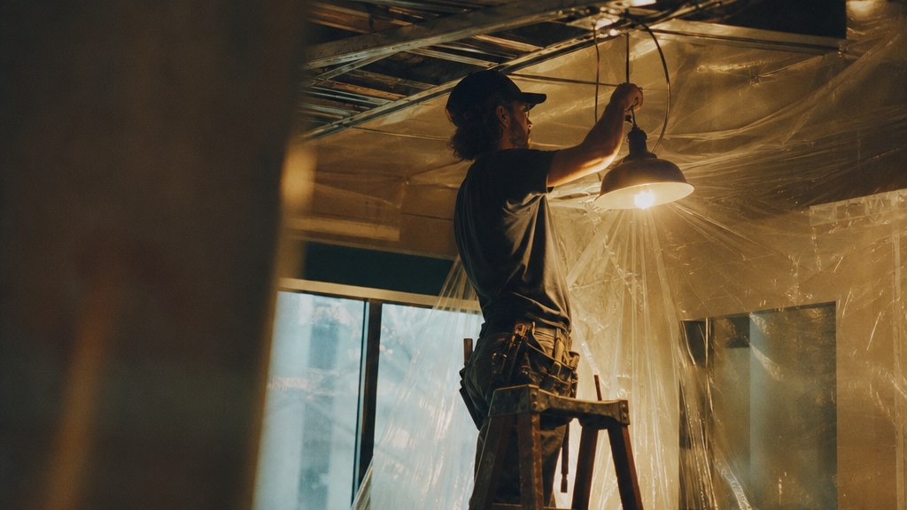 Electrician near the top of a ladder steadying a pendant fixture in a half-wired ceiling under dust sheeting