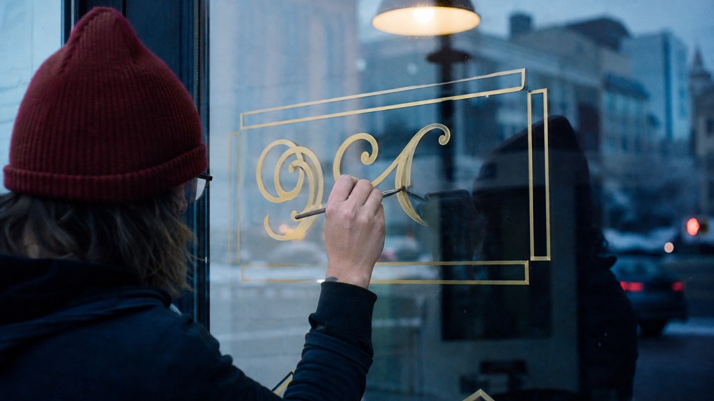 A sign painter finishing gold lettering on a shopfront window from the street side, photographed in Wichita