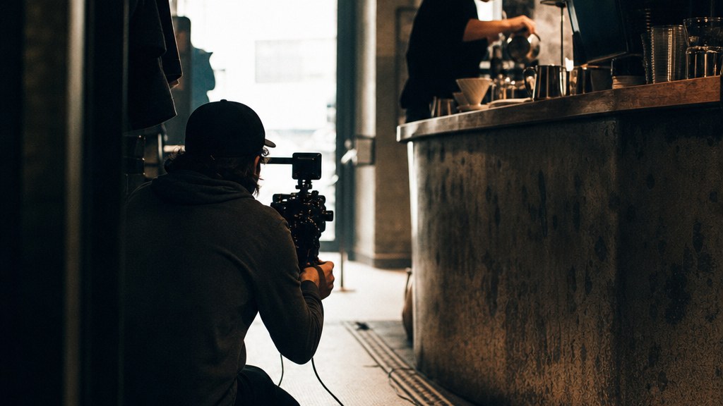 Camera operator crouched low filming a barista mid-pour behind a counter, shop lights blown out behind them