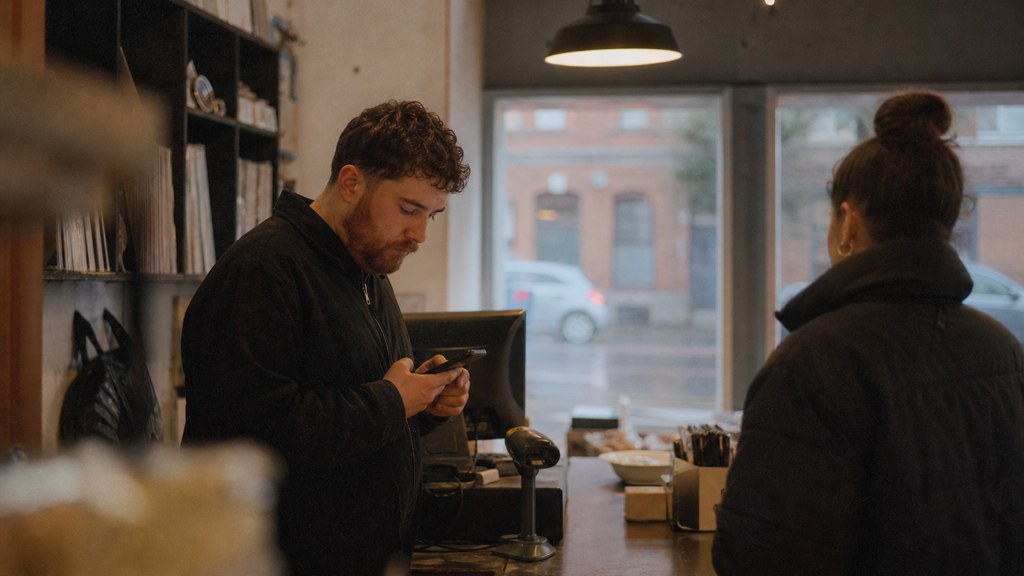A shop manager standing behind the counter checking a phone while a customer waits at the till, photographed in Belfast
