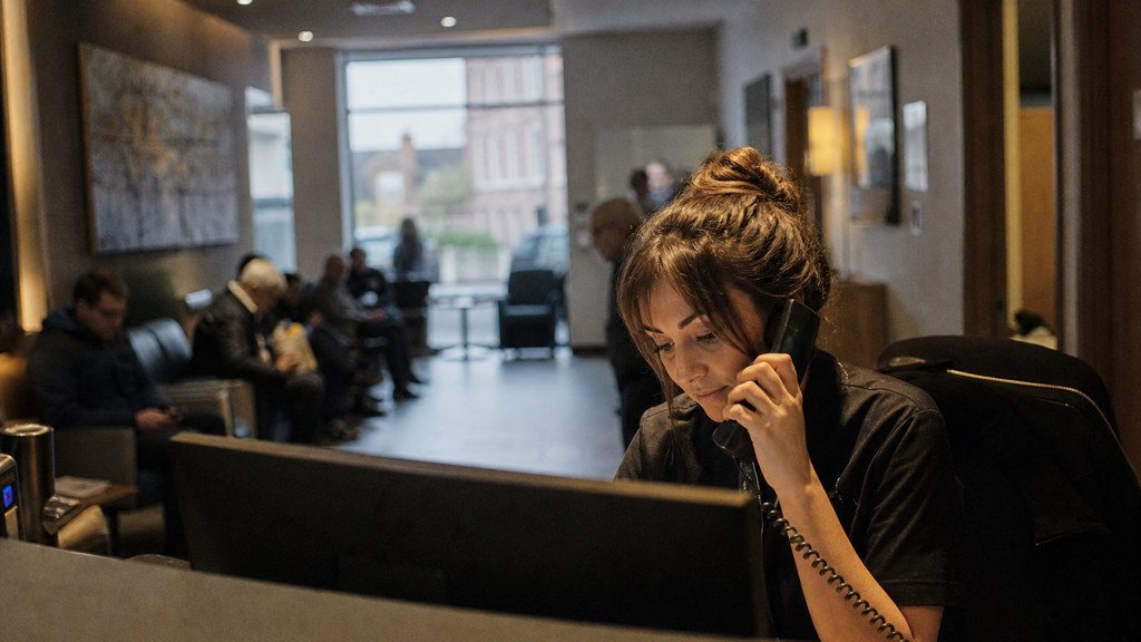 A clinic receptionist taking a phone call at a busy front desk with patients waiting, photographed in Belfast