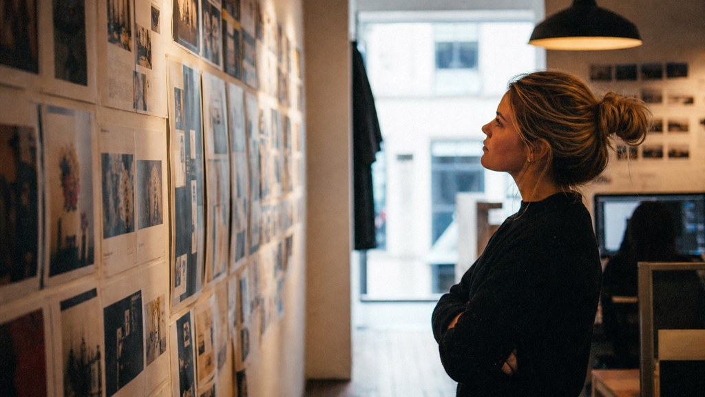 A media planner standing at a wall of printed layouts in a narrow agency corridor, photographed in Belfast