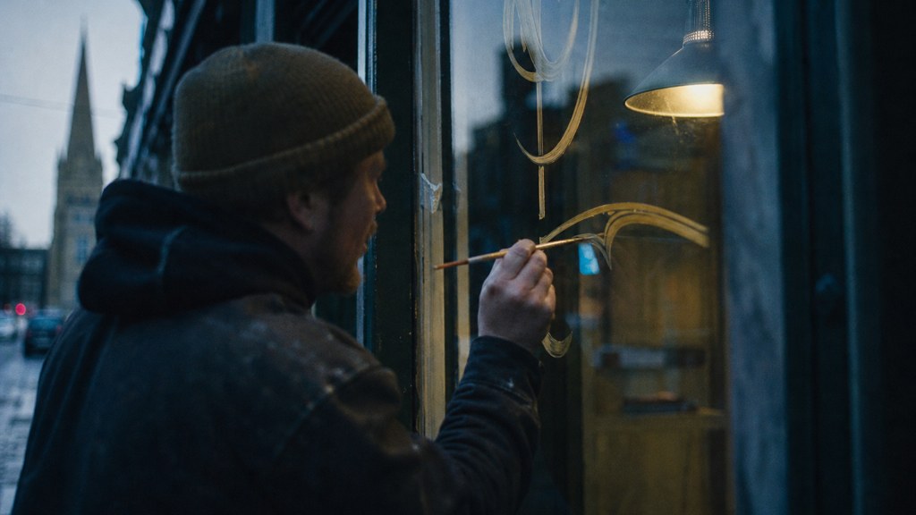 A sign painter finishing gold lettering on a shopfront window from the street side, photographed in Belfast