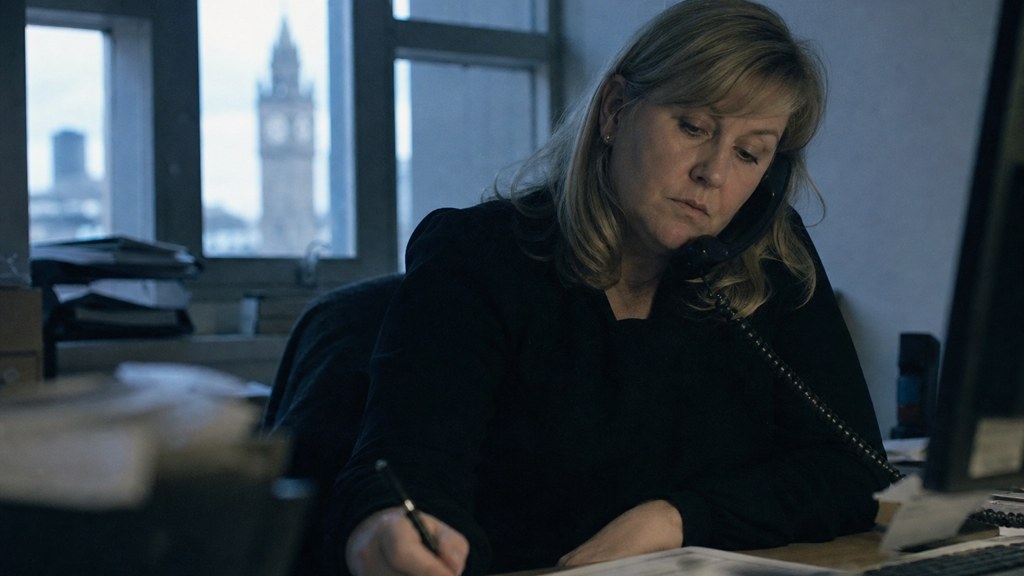 An office manager at a cluttered desk with a landline handset tucked under one ear, photographed in Belfast