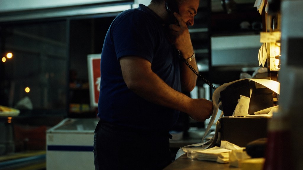 Franchise owner answering a ringing phone at a service counter late at night under a flickering fluorescent tube