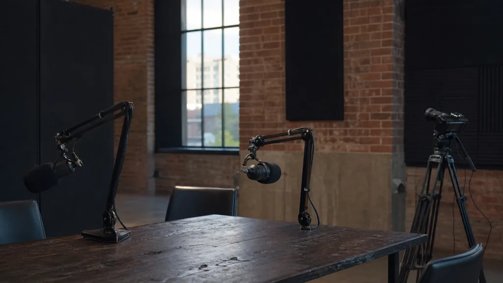 Editorial photograph of an empty podcast studio in a Spokane Washington brick warehouse conversion with two microphones on boom arms, a camera on a tripod and dark acoustic panels, lit by cool morning light through tall industrial windows