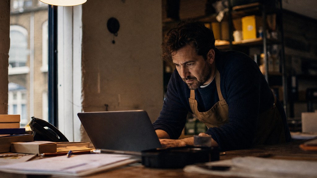 A small-business owner in an apron leaning over a laptop on a cluttered back-office desk late in the day, photographed in Bristol