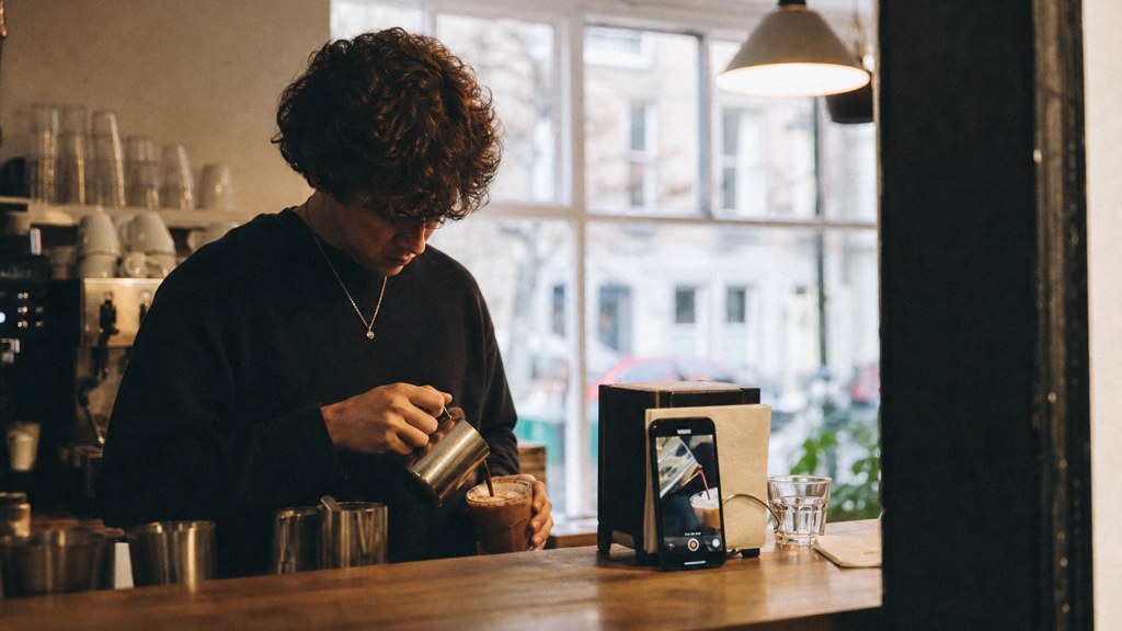 A young cafe worker filming a drink being poured on a phone propped against a napkin holder, photographed in Bristol