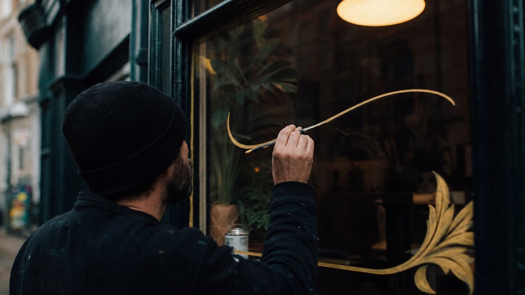 A sign painter finishing gold lettering on a shopfront window from the street side, photographed in Bristol