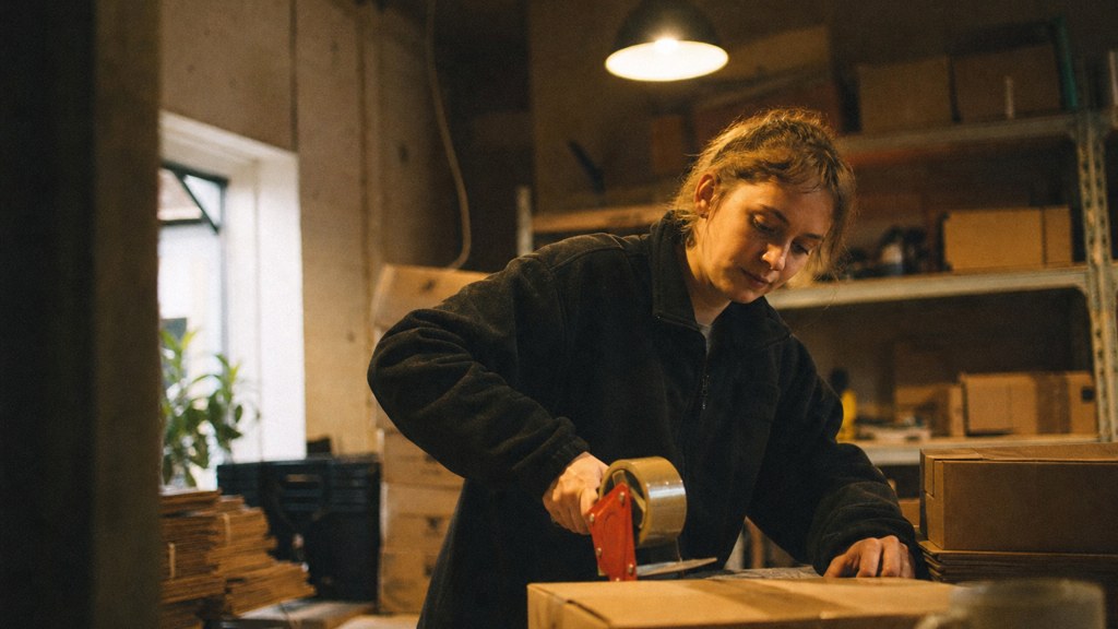 A warehouse worker taping shipping boxes under strip lighting in a small fulfilment room, photographed in Bristol