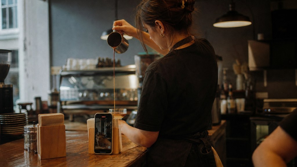 A young cafe worker filming a drink being poured on a phone propped against a napkin holder, photographed in Cardiff