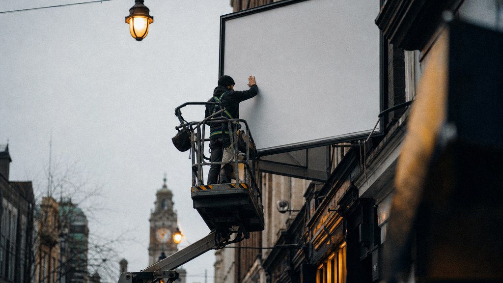 A crew member on a lift installing a blank billboard panel above a busy street, photographed in Cardiff