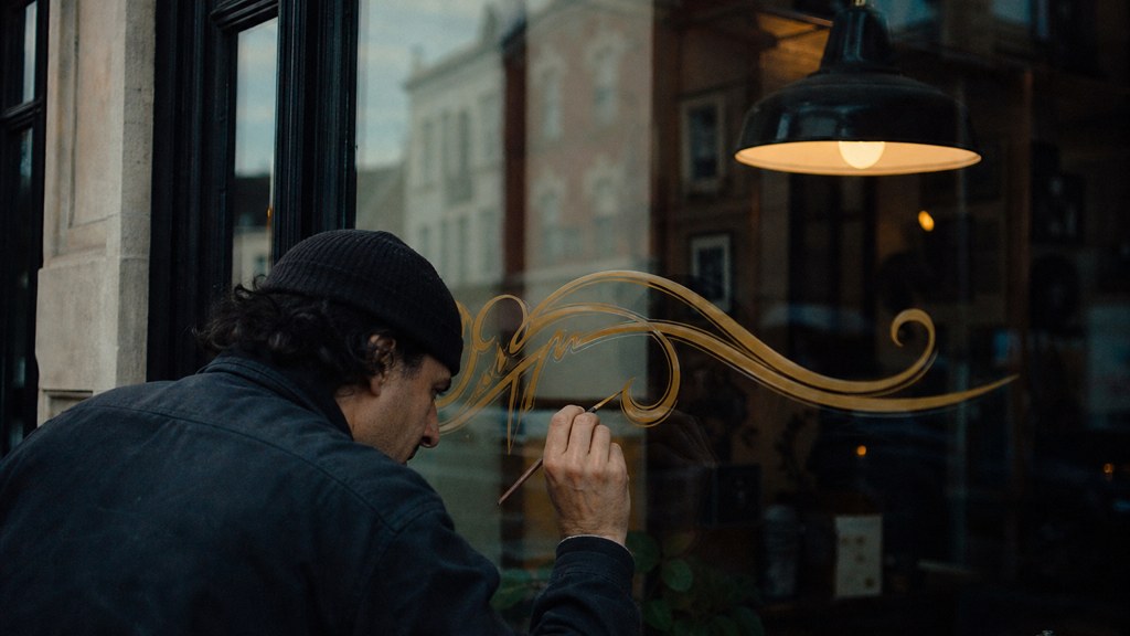A sign painter finishing gold lettering on a shopfront window from the street side, photographed in Cardiff