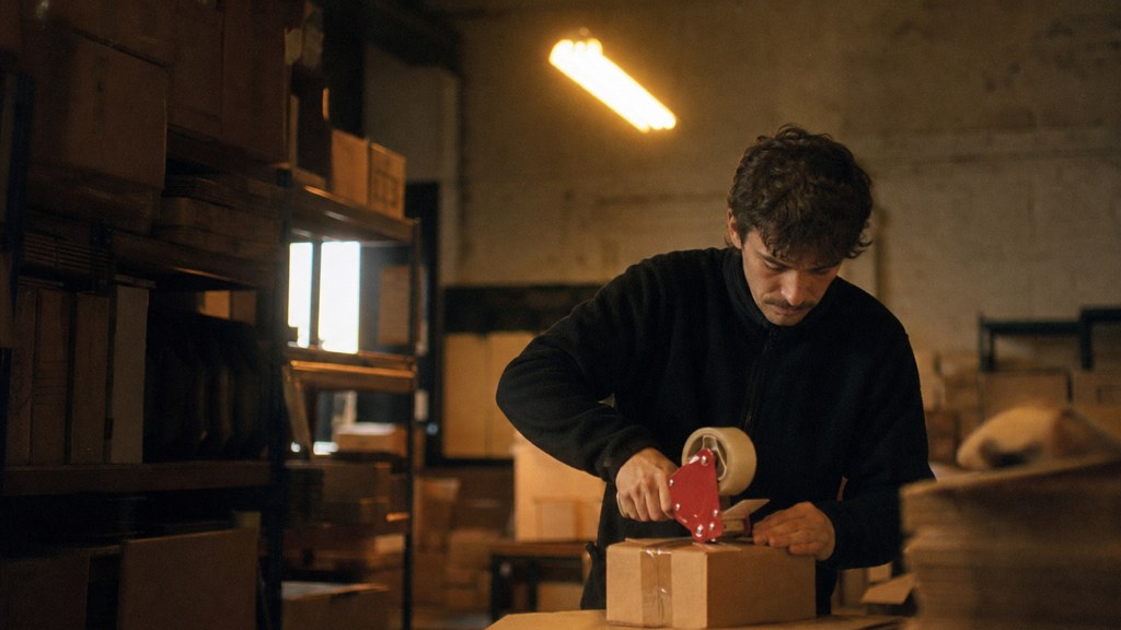 A warehouse worker taping shipping boxes under strip lighting in a small fulfilment room, photographed in Cardiff