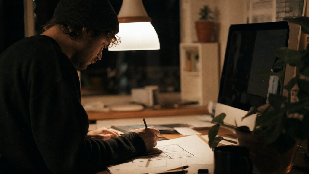 A studio designer sketching a page layout on paper beside a monitor in a dim room, photographed in Cardiff