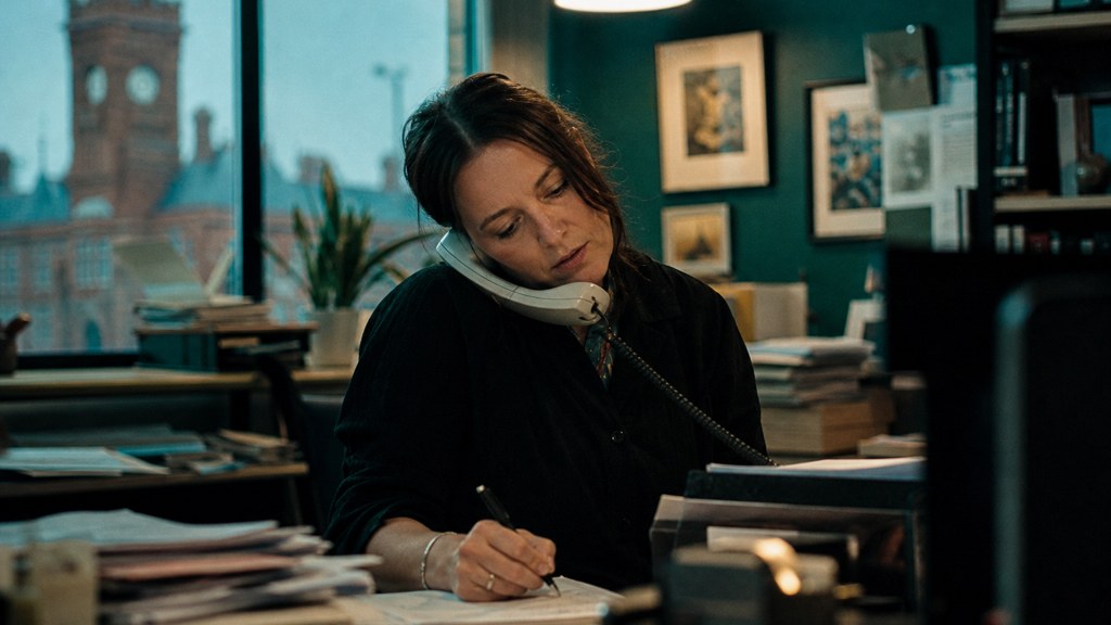 An office manager at a cluttered desk with a landline handset tucked under one ear, photographed in Cardiff