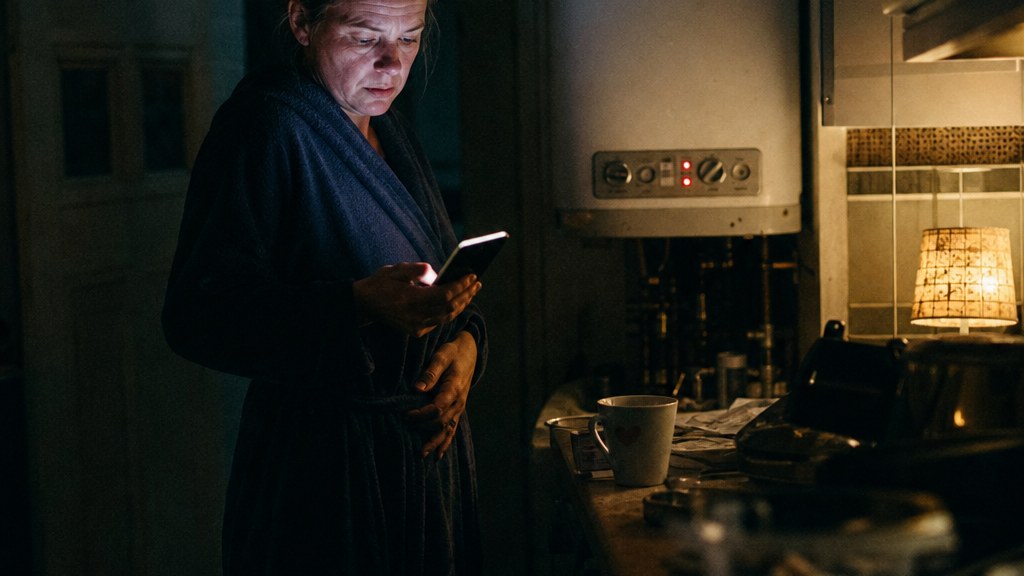 Homeowner in a cold kitchen at night holding a phone while a boiler panel flashes an error behind her