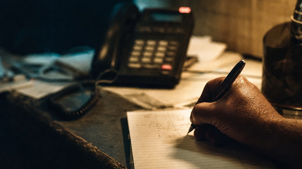 Office worker at a small contractor desk writing a caller's answer onto a notepad beside a ringing phone