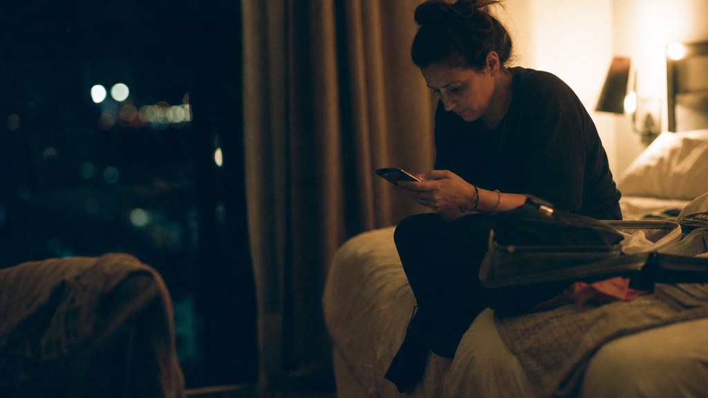 Traveller sitting on the end of a hotel bed at night comparing properties on a phone with a half-packed suitcase open beside her